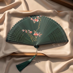 May include: A dark green folding hand fan featuring a delicate painted branch with pink and white blossoms. The fan has intricate cut-out patterns along the sides and a matching dark green tassel attached to the base with a small green bead.