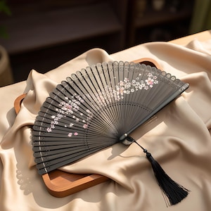 May include: A black folding hand fan featuring a delicate white and pink cherry blossom pattern across the ribs. The top edge of the fan has a scalloped, laser-cut design, and a long black tassel is attached to the base pivot point.