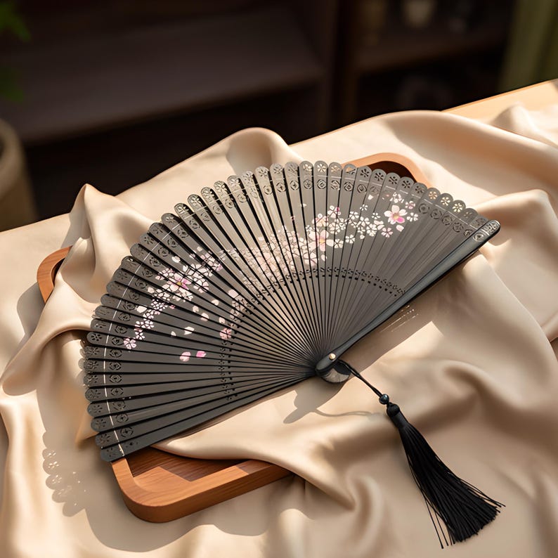 May include: A black folding hand fan featuring a delicate white and pink cherry blossom pattern across the ribs. The top edge of the fan has a scalloped, laser-cut design, and a long black tassel is attached to the base pivot point.