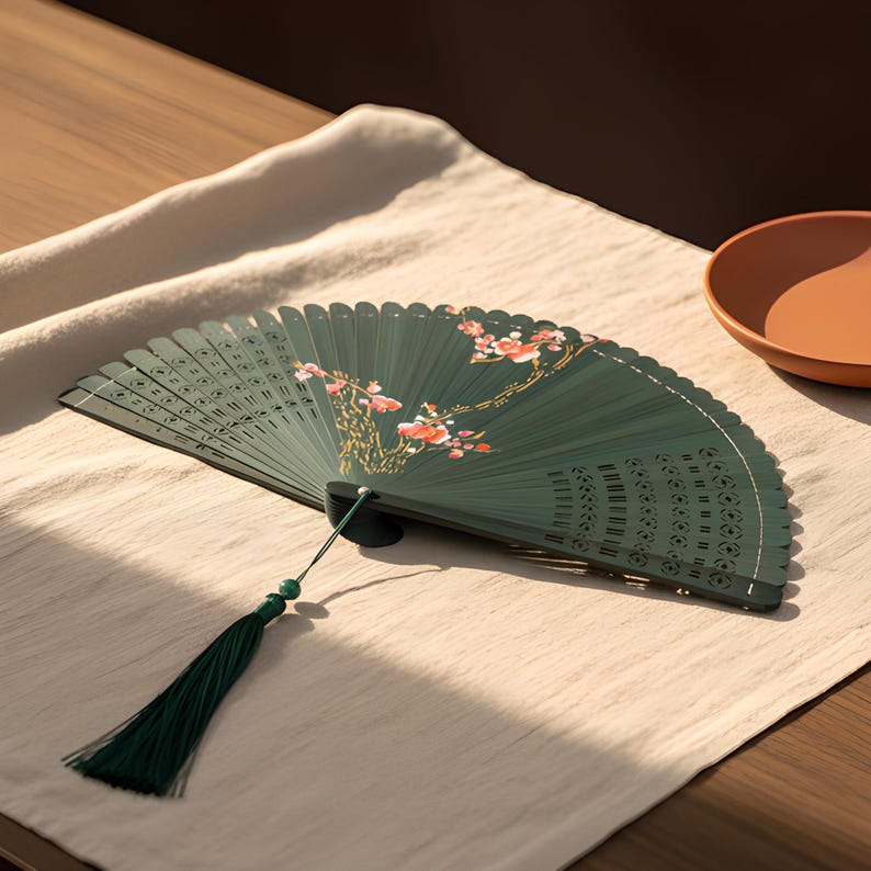May include: A dark green folding hand fan featuring a delicate floral pattern with pink and white blossoms on the surface. The fan includes intricate cut-out details along the edges and a long, dark green decorative tassel attached at the base.