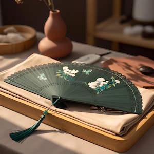 May include: A dark green folding hand fan featuring a white floral pattern and a small green butterfly illustration. The fan has a decorative laser-cut edge and a long green tassel attached to the handle with a small green bead accent.