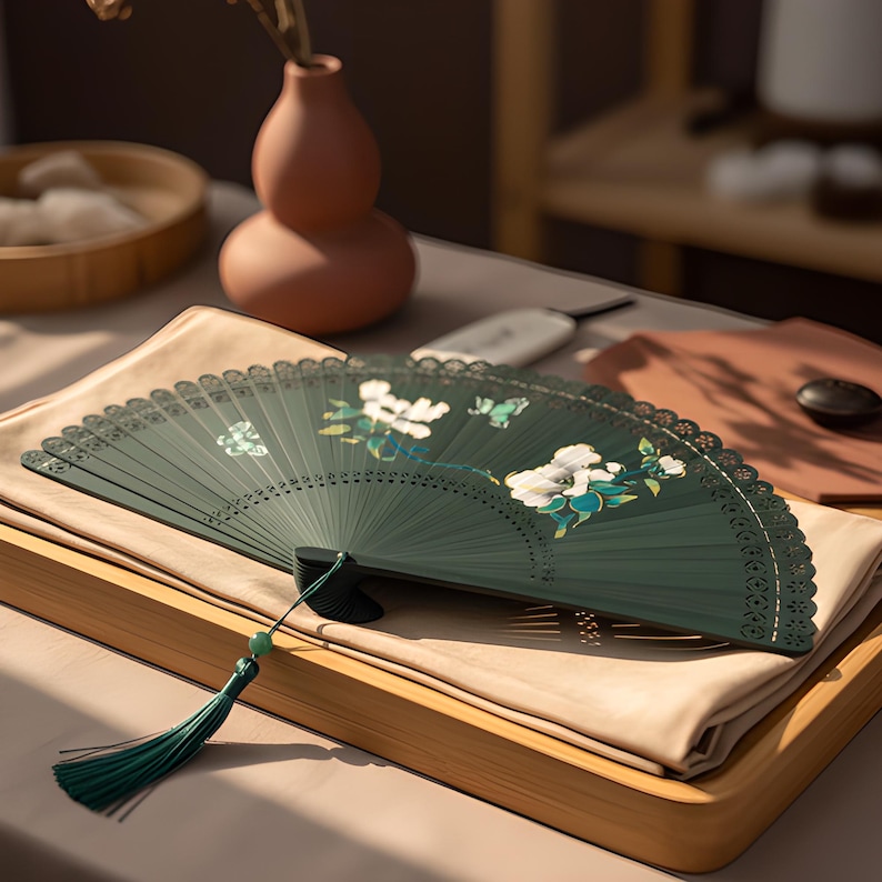 May include: A dark green folding hand fan featuring a white floral pattern and a small green butterfly illustration. The fan has a decorative laser-cut edge and a long green tassel attached to the handle with a small green bead accent.