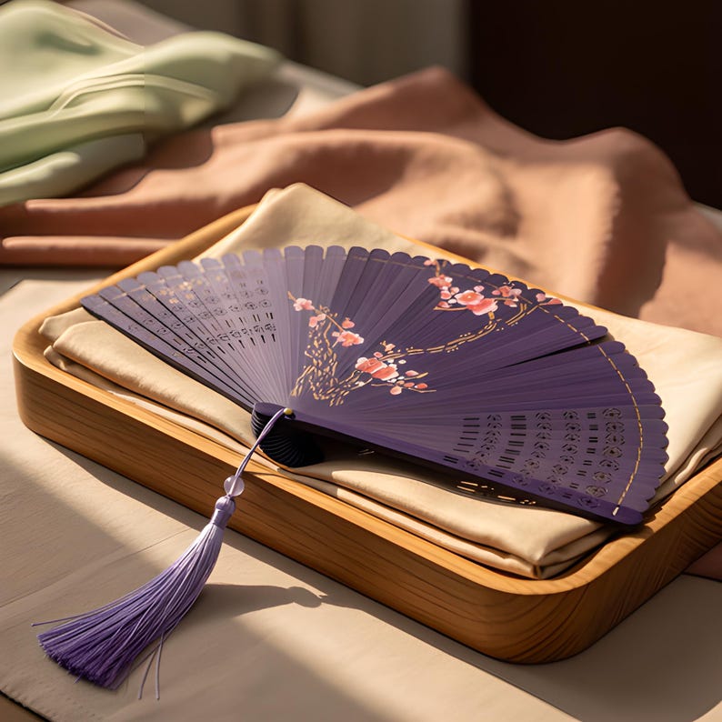 May include: A deep purple folding hand fan featuring a delicate pink and white floral branch design. The fan includes intricate cut-out patterns along the ribs and is finished with a long, light purple tassel attached to the base pivot point.