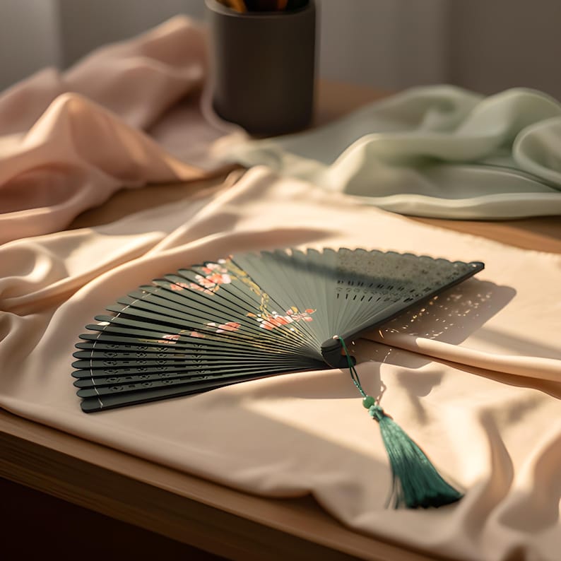 May include: A dark green folding hand fan featuring a delicate floral pattern with pink and white blossoms. The fan has a series of thin, parallel slats and a decorative dark green tassel attached to the base with a small green bead accent.