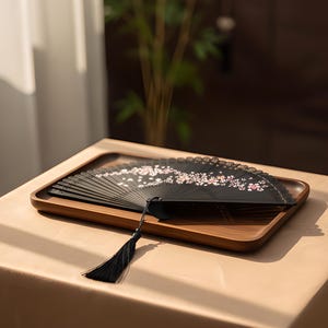 May include: A black folding hand fan featuring a delicate pink and white cherry blossom floral pattern across the ribs. The fan includes a long black tassel attached to the handle, showcasing intricate cut-out details along the upper edge.