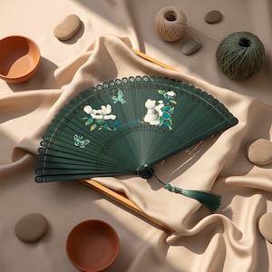 May include: A dark green folding hand fan featuring white floral and butterfly illustrations, intricate laser-cut patterns along the top edge, and a matching green tassel attached to the base with a small green bead, creating a decorative accessory.