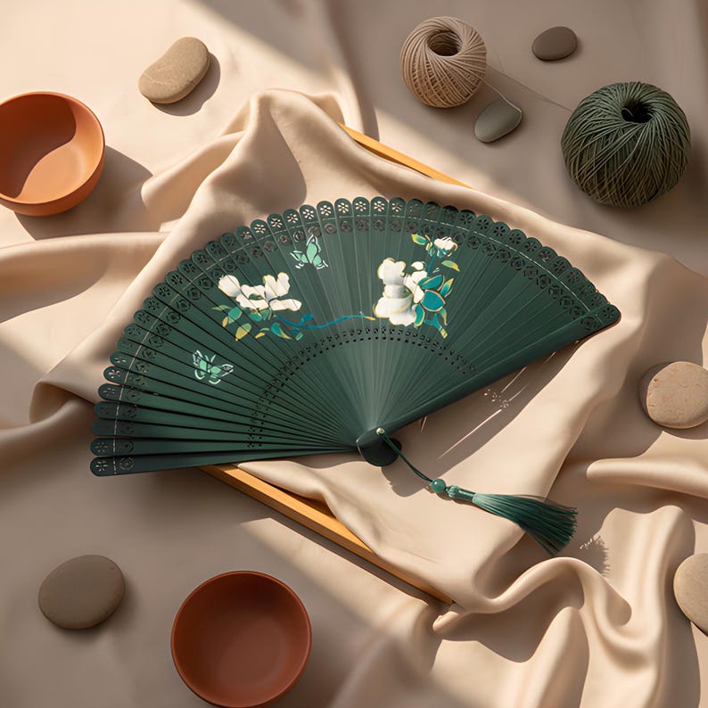 May include: A dark green folding hand fan featuring white floral and butterfly illustrations, intricate laser-cut patterns along the top edge, and a matching green tassel attached to the base with a small green bead, creating a decorative accessory.