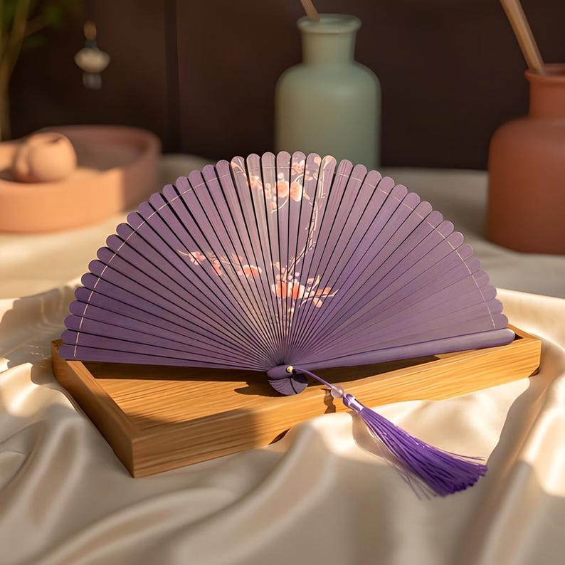 May include: A purple folding hand fan featuring a delicate floral branch pattern with light pink blossoms. The fan is constructed with thin, evenly spaced ribs and includes a matching purple tassel attached to the base with a small bead.