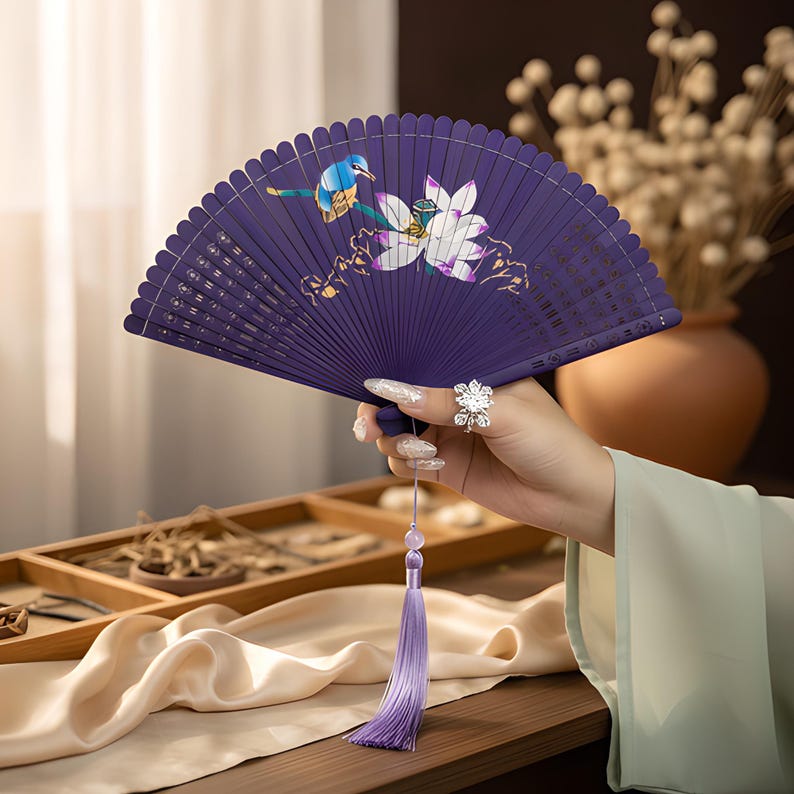 May include: A deep purple folding hand fan featuring a painted design of a small blue bird perched near a white lotus flower. The fan includes a long, light purple silk tassel hanging from the base, attached by a small, light-colored bead.