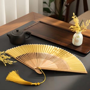 May include: A gold and tan folding fan with a yellow tassel is open on a dark surface. The fan is made of paper and wood. A small black teapot, a white vase with yellow flowers, and a wooden tray are in the background.