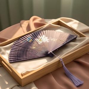 May include: A folding hand fan featuring a purple gradient background with a white floral design and a small blue bird illustration. The fan has a purple tassel attached to the handle, which is secured with a small metal pin at the pivot point.