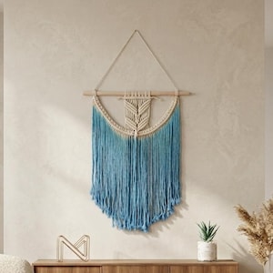 Könnte beinhalten: Makramee-Wandbehang mit blauem Farbverlauf und Holzstab. Der Wandschmuck hängt über einem Holzschrank. Eine kleine Topfpflanze und eine dekorative Vase sind ebenfalls zu sehen.