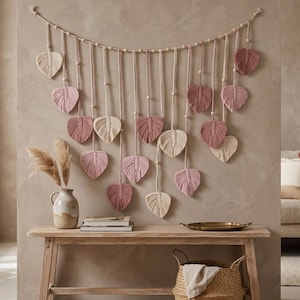 May include: A macrame wall hanging featuring a horizontal cord adorned with wooden beads and multiple dangling leaf-shaped fiber elements in shades of cream, dusty rose, and mauve, suspended in a symmetrical arrangement against a neutral wall.