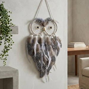 May include: An owl-shaped dreamcatcher with a beige frame and white web design. The owl's eyes are black, and the body is made of gray and white speckled feathers. The dreamcatcher hangs from a string.