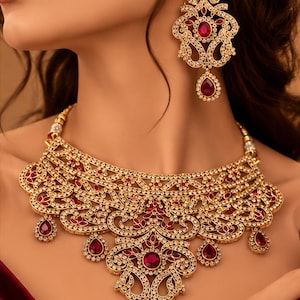 Puede incluir: Un conjunto de collar y pendientes en tono dorado con intrincados trabajos de metal en filigrana, incrustados con pequeños cristales transparentes y acentuados por piedras de color rojo intenso en forma de pera y corte ovalado.