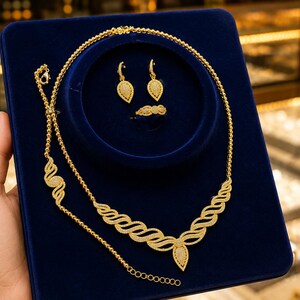 May include: A gold jewelry set featuring a necklace with a wavy, textured pendant, a matching bracelet, a pair of teardrop-shaped dangle earrings, and a ring. The pieces are displayed on a dark blue velvet board, showcasing intricate gold metalwork.