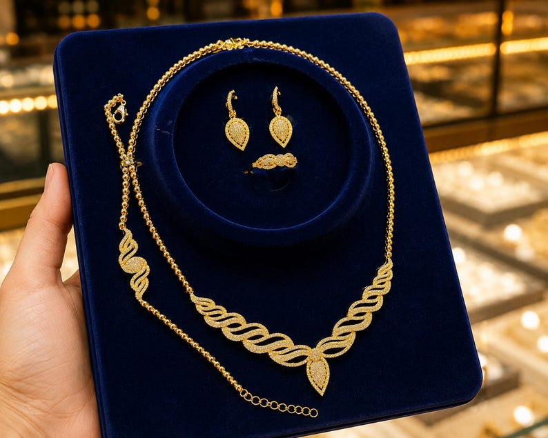 May include: A gold jewelry set featuring a necklace with a wavy, textured pendant, a matching bracelet, a pair of teardrop-shaped dangle earrings, and a ring. The pieces are displayed on a dark blue velvet board, showcasing intricate gold metalwork.