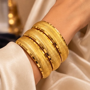 May include: A set of three wide gold bangles featuring a brushed, textured finish on the surface and polished, smooth, reflective edges. The bracelets are stacked together, showcasing a consistent metallic yellow colour and a rounded, convex profile.
