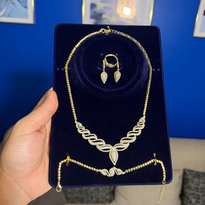 May include: A gold-toned jewelry set displayed on a dark blue velvet board, featuring a necklace with a central teardrop pendant, a matching bracelet, and a ring with two teardrop-shaped accents, all embellished with small clear stones.