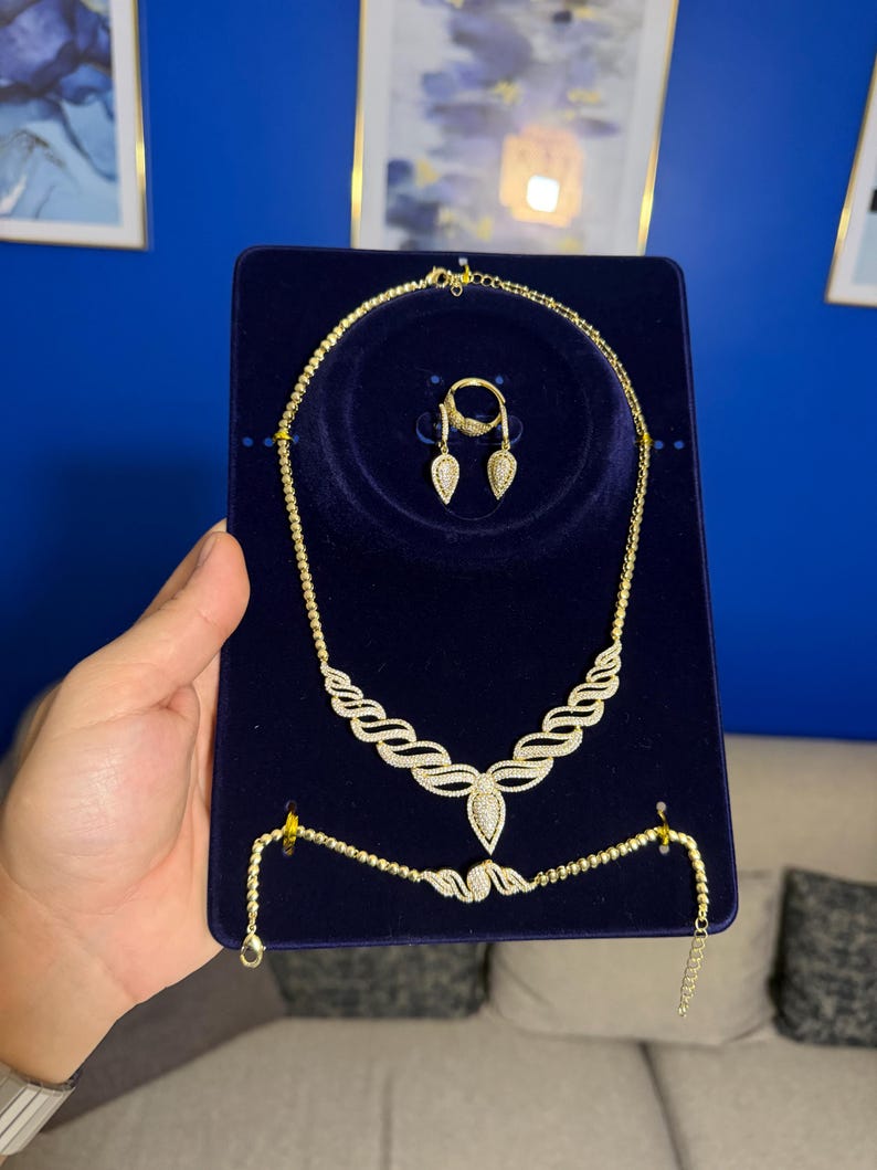 May include: A gold-toned jewelry set displayed on a dark blue velvet board, featuring a necklace with a central teardrop pendant, a matching bracelet, and a ring with two teardrop-shaped accents, all embellished with small clear stones.