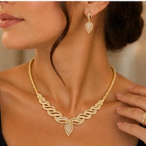 May include: A gold-toned jewelry set consisting of a necklace, drop earrings, and a ring. The necklace features a central teardrop pendant with a pav&eacute;-set surface, flanked by wavy, textured links. The matching earrings and ring mirror the teardrop design.