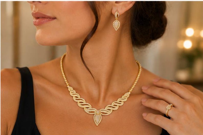 May include: A gold-toned jewelry set consisting of a necklace, drop earrings, and a ring. The necklace features a central teardrop pendant with a pav&eacute;-set surface, flanked by wavy, textured links. The matching earrings and ring mirror the teardrop design.