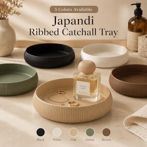 Könnte beinhalten: Ein Set aus fünf runden, gerippten Ablageschalen in Schwarz, Weiß, Eiche, Grün und Braun. Der Text "5 Colors Available" und "Japandi Ribbed Catchall Tray" steht darüber. Eine Schale enthält ein Parfümfläschchen, eine Goldkette und einen Ring.