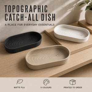 May include: Three oval catch-all dishes featuring a topographic map pattern on the interior base. The dishes are presented in black, white, and beige colors. Text on the image reads: TOPOGRAPHIC CATCH-ALL DISH, A PLACE FOR EVERYDAY ESSENTIALS.