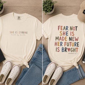 以下が含まれることがあります： インスピレーションを与える引用句が書かれたクリーム色のTシャツ2枚。前面のシャツには、カラフルな文字で「She is Strong, built by faith」と書かれています。背面のシャツには、ハートと「Jeremiah 29:1」とともに「Fear Not, She is Made New, Her Future is Bright」と書かれています。