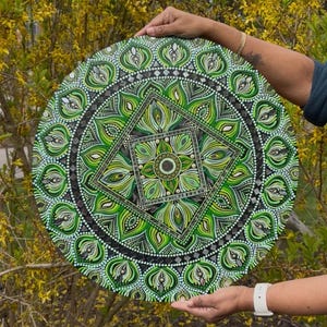 May include: A round, hand-painted mandala with a complex geometric design. The artwork features shades of green, black, and white, with a central square and circular patterns. The piece is held up against a backdrop of yellow flowers.
