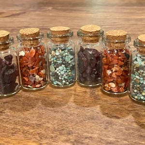 Könnte beinhalten: Sechs kleine, durchsichtige Glasflaschen mit Korkstopfen, die jeweils mit verschiedenen Arten bunter Mineralsteine gefüllt sind. Die Sammlung umfasst Gläser mit dunkelvioletten, leuchtend orangefarbenen und türkisfarbenen Steinsplittern und kleinen Kieselsteinen.