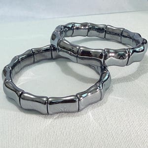 Könnte beinhalten: Zwei silberfarbene Armbänder. Jedes Armband besteht aus rechteckigen, gebogenen Segmenten, die wie Bambus aussehen. Die Armbänder liegen auf einer weißen Oberfläche, mit weicher Beleuchtung.
