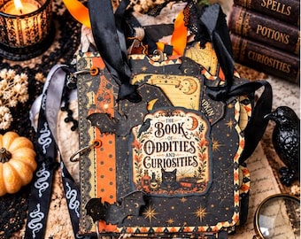 Könnte beinhalten: Ein handgefertigtes Sammelalbum mit dem Titel The Book of Oddities and Curiosities, verziert mit schwarz-orangem Musterpapier, ausgestanzten Fledermäusen und schwarzen Satinbändern. Das Cover zeigt ein Etikett mit einer schwarzen Katze.