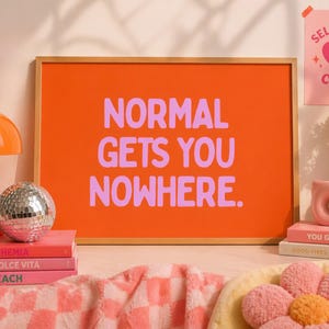 May include: An orange framed print with the words "NORMAL GETS YOU NOWHERE" in lavender. The image includes a disco ball, a stack of pink books, a pink donut-shaped vase, and a "SELF LOVE CLUB" poster.