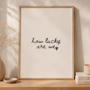 Op de afbeelding: Een ingelijste print met de tekst "how lucky are we" in zwart schrift. De print wordt getoond op een houten oppervlak, samen met een vaas met gedroogde bloemen, een kleine kom, een kaars en twee boeken.