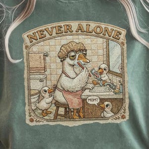 Może przedstawiać: Szałwiowa zielona koszulka z zabawną ilustracją gęsi w łazience z kaczątkami, nad ilustracją znajduje się napis "NEVER ALONE". Gęś ma na sobie czepek kąpielowy i fartuch. Koszulka jest w stylu casual.