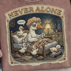 Może przedstawiać: Bluza w kolorze mauve z zabawną ilustracją. Przedstawia gęś w łóżku, otoczoną przez kaczątka, z napisem "NEVER ALONE" u góry. Gęś trzyma butelkę, z napisem "MOM?" w dymku.
