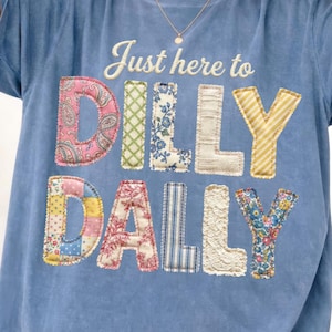 Könnte beinhalten: Ein blaues T-Shirt mit dem Aufdruck "Just here to Dilly Dally" im Patchwork-Stil. Die Buchstaben bestehen aus verschiedenen gemusterten Stoffen, darunter Paisley-, Blumen- und Streifenmuster. Das Shirt hat kurze Ärmel.