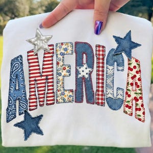 Op de afbeelding: Witte sweatshirt met het woord "AMERICA" in patchwork. De letters zijn gemaakt van denim, rood-witte strepen, bloemenstof en kersenprint. Er zijn ook verschillende stervormige applicaties op de sweatshirt genaaid.