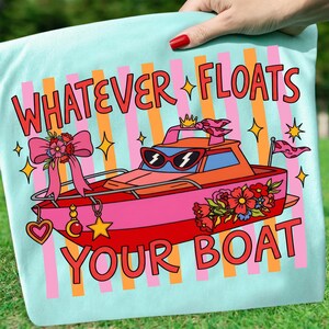 May include: A light blue t-shirt featuring a graphic of a pink and orange boat wearing sunglasses. The text reads "WHATEVER FLOATS YOUR BOAT" in red letters. The design includes a large pink bow, floral accents, stars, and vertical stripes.