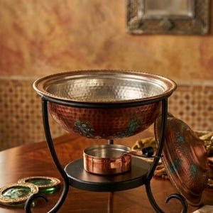 May include: Copper fondue pot with a hammered texture, resting on a black metal stand. The pot has a matching lid and a small copper pot below. Includes three green and gold coasters. A decorative piece for entertaining.