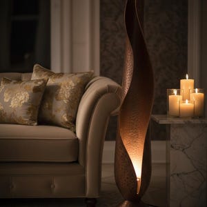 Op de afbeelding: Een hoge, koperkleurige vloerlamp met een uniek, gebogen ontwerp. De lamp heeft een getextureerd oppervlak en straalt een warme gloed uit. De lamp staat naast een beige bank met bloemenkussens en een marmeren bijzettafel met kaarsen.