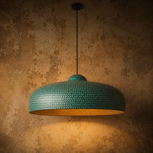 Könnte beinhalten: Eine türkisfarbene Pendelleuchte mit strukturierter, genoppter Oberfläche und goldfarbenem Innenraum. Die Leuchte hängt an einem schwarzen Kabel und einer Deckenhalterung. Das Design der Lampe erinnert an eine gehämmerten Metalloberfläche.
