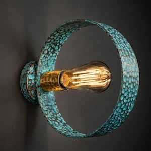 Op de afbeelding: Decoratieve wandlamp met een turquoise en bronzen afwerking. De lamp heeft een cirkelvormig ontwerp met een textuurpatroon en een vintage gloeilamp. Gemonteerd op een donkere muur.