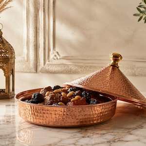 Könnte beinhalten: Ein Kupfer-Tajine mit gehämmerten Texturen, teilweise geöffnet, um einen Eintopf mit Trockenfrüchten und Nüssen zu zeigen. Das Tajine hat einen konischen Deckel mit einem goldfarbenen Knauf. Eine dekorative Laterne und eine Vase befinden sich im Hintergrund.
