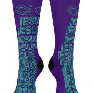 Könnte beinhalten: Ein Paar violette Crew-Socken mit einem sich wiederholenden vertikalen Muster des Wortes JESUS in hellgrüner Schrift. Oben auf jeder Socke befindet sich ein hellgrüner Umriss eines Fischsymbols. Die Socken haben einfarbige schwarze Zehen- und Fersenbereiche.