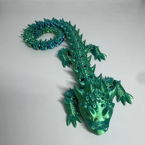 Op de afbeelding: Een 3D-geprint gearticuleerd draakspeelgoed in groen- en blauwtinten. De draak heeft een gedetailleerd, getextureerd oppervlak met spikes langs zijn rug en staart. De kop van de draak is zichtbaar, met gedetailleerde ogen en een licht geopende mond.