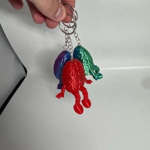 Può includere: Tre portachiavi con figure uniche e colorate stampate in 3D. I portachiavi presentano una figura rossa con braccia e gambe, una figura viola e blu e una figura verde. Ogni figura è attaccata a un portachiavi argentato.