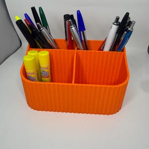 Puede incluir: Un organizador de escritorio naranja con múltiples compartimentos, lleno de bolígrafos, rotuladores y barras de pegamento. El organizador tiene una textura acanalada y está diseñado para guardar varios utensilios de escritura y material de oficina. La imagen muestra varios bolígrafos y barras de pegamento.
