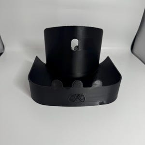 May include: A black 3D-printed object with a curved base and a cylindrical top. The object has a small hole near the top and a decorative emblem on the front. The design suggests a holder or stand.